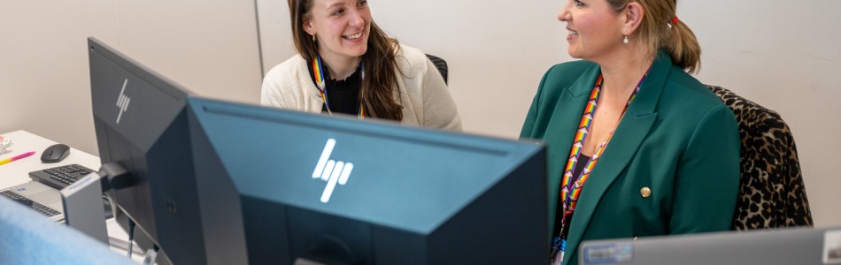 Photo of two female staff members sat behind computers smiling, in an office based scenario.