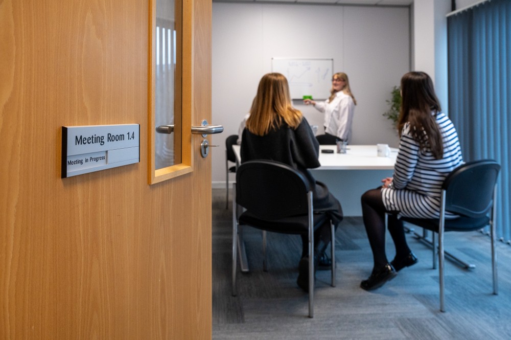 Three women sat in a meeting room with whiteboard, chairs and table